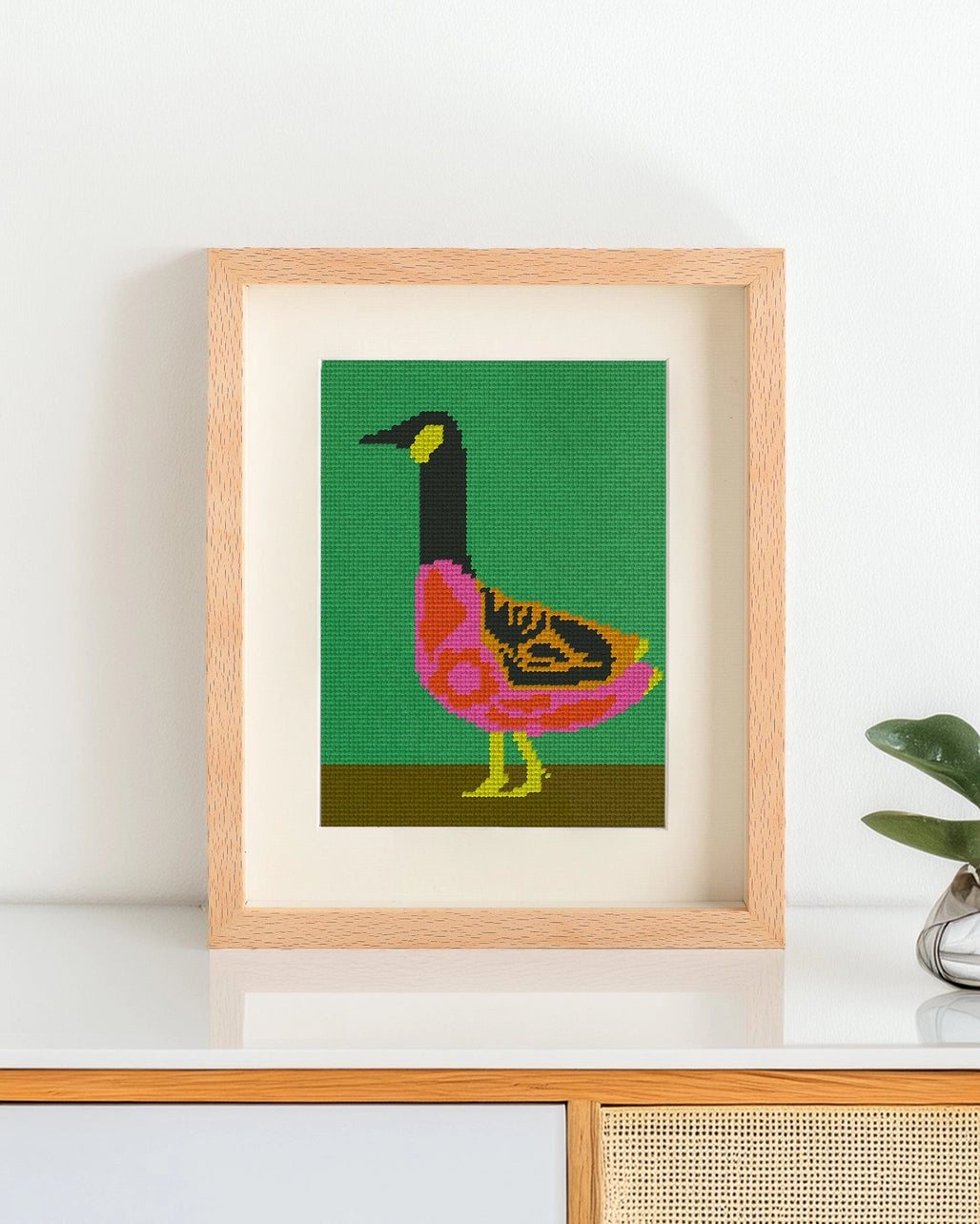 Framed needlepoint artwork with a colorful goose design resting on a white table against a white background