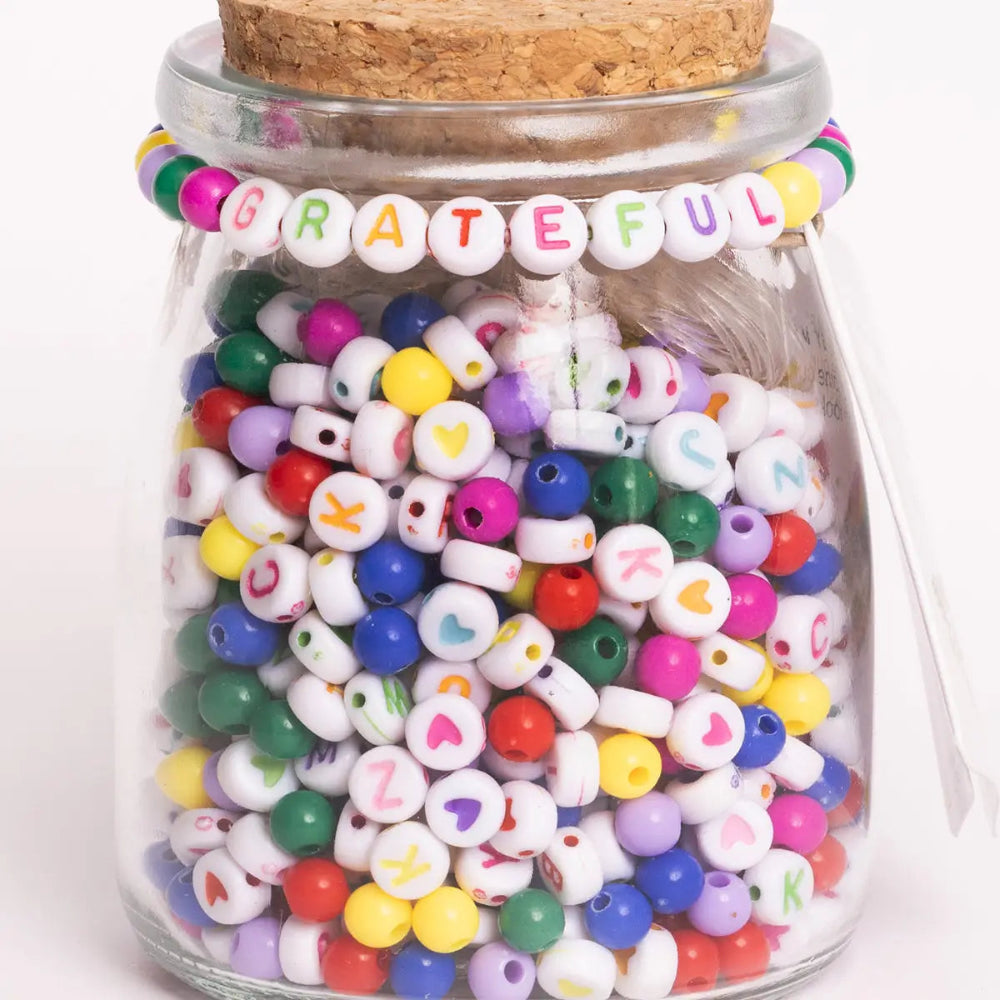 A glass jar with  different types of beads, including colorful alphabet beads and solid color beads. There is a beaded bracelet around the top with beads that spell "grateful" in the center. 