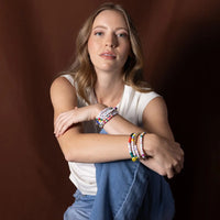 A person with a white shirt and jeans is sitting with one leg up and their arms embracing their leg. They have multiple beaded bracelets on their arm. 