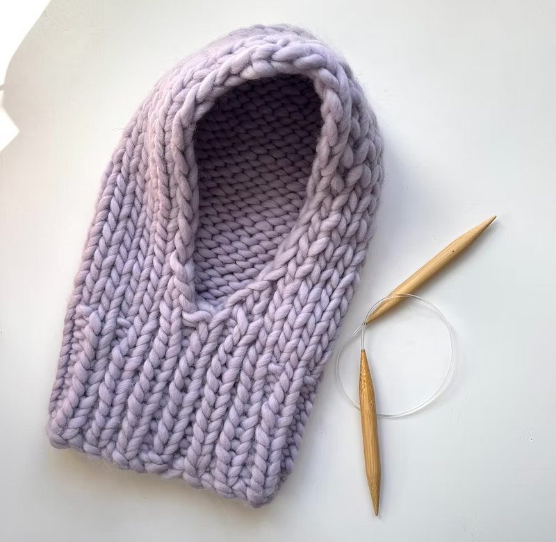Liliac knit hood on a white background next to wooden circular knitting needles.