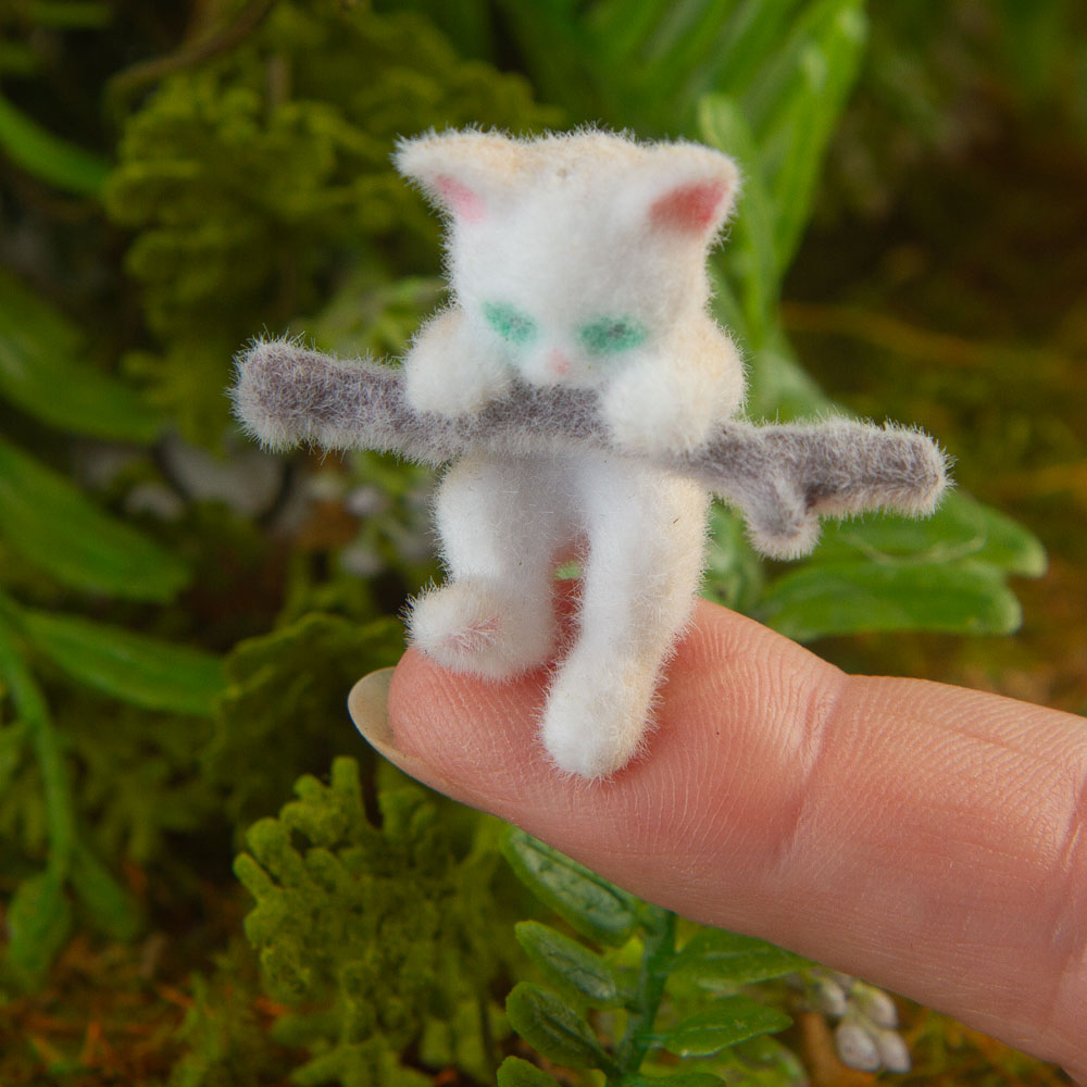 A small white fuzzy cat figurine with mint-green eyes and pink inner ears, holding a light brown branch, resting on a human finger against a blurred green plant background.