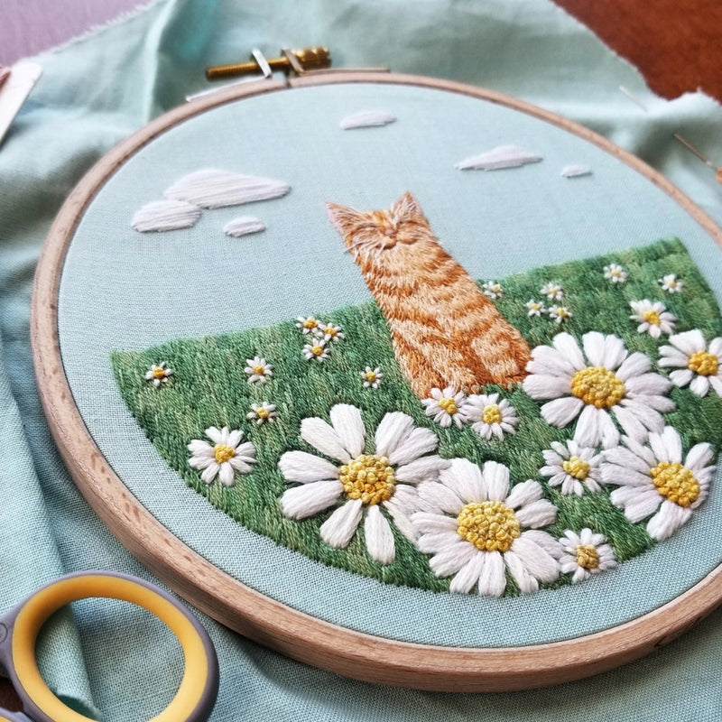 Embroidery hoop with a detailed design of an orange cat in a flower field