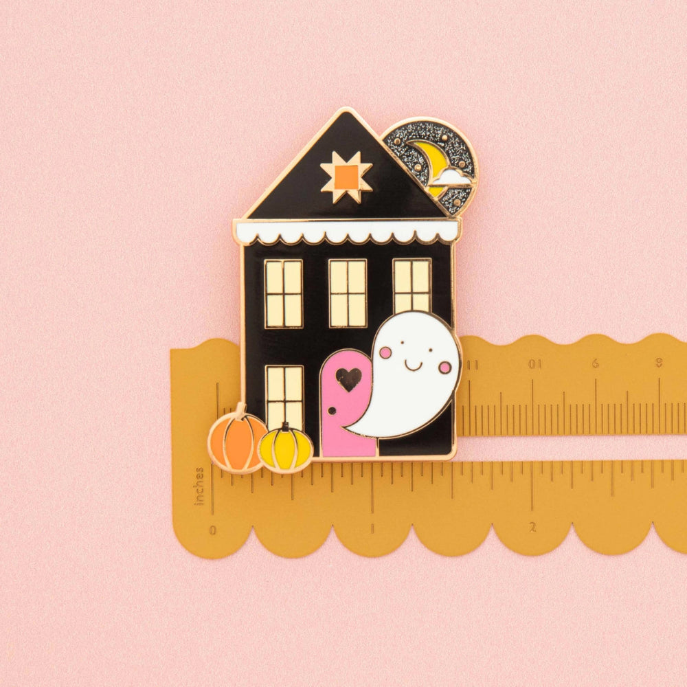 A cute Halloween-themed enamel pin featuring a black haunted house, a smiling white ghost, pumpkins, and a crescent moon, shown next to a yellow scalloped-edge ruler on a soft pink background.