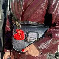 Person holding a black handbag with a red heart-shaped keychain, wearing a maroon leather jacket.