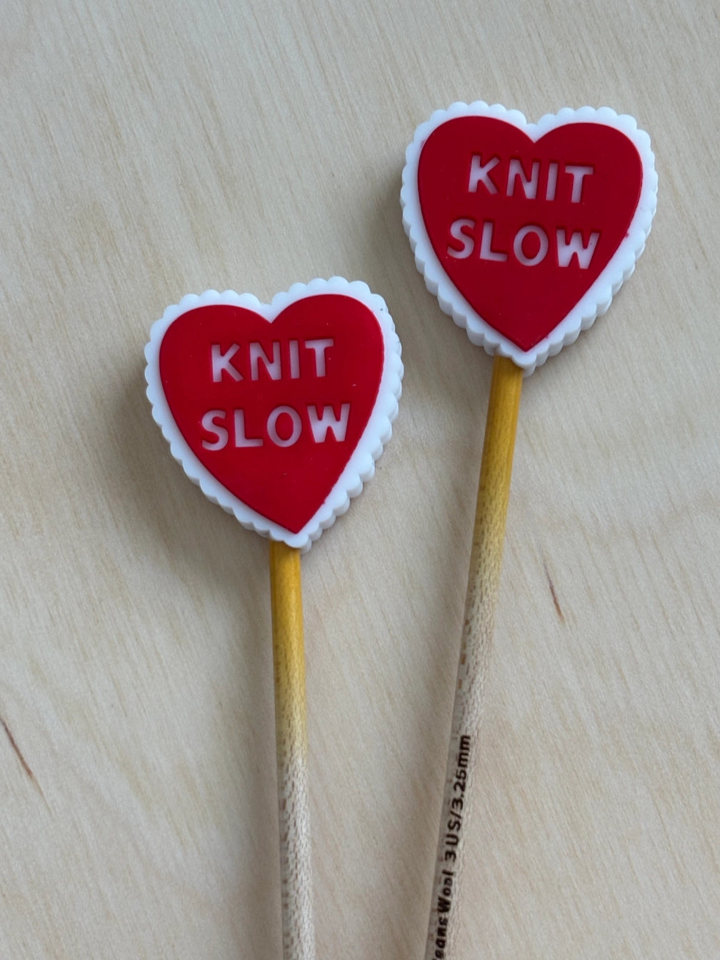 Two heart-shaped knitting needle tips with 'KNIT SLOW' text on a wooden surface.
