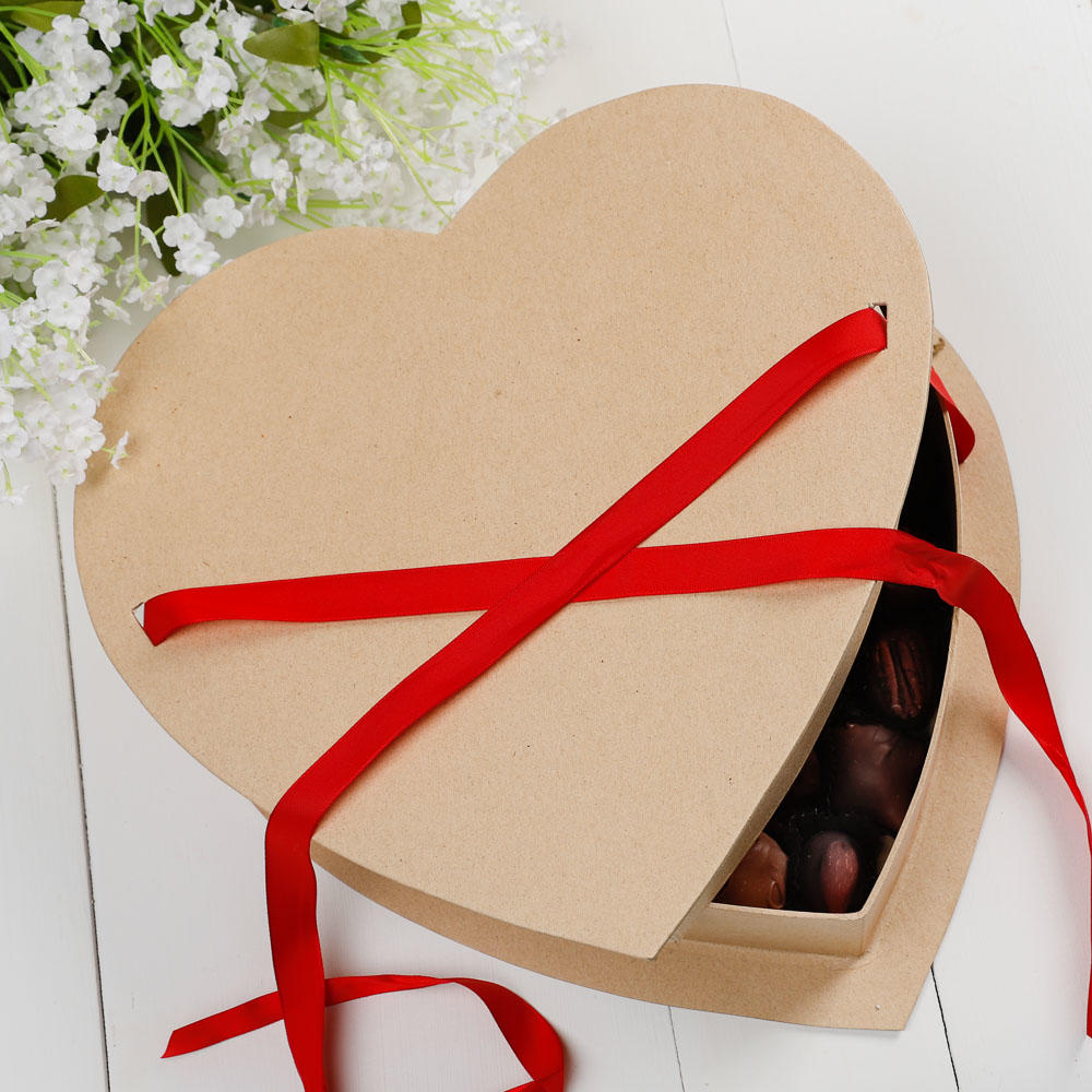 Heart-shaped cardboard gift box with red ribbon and chocolates inside on a white surface with flowers in the background