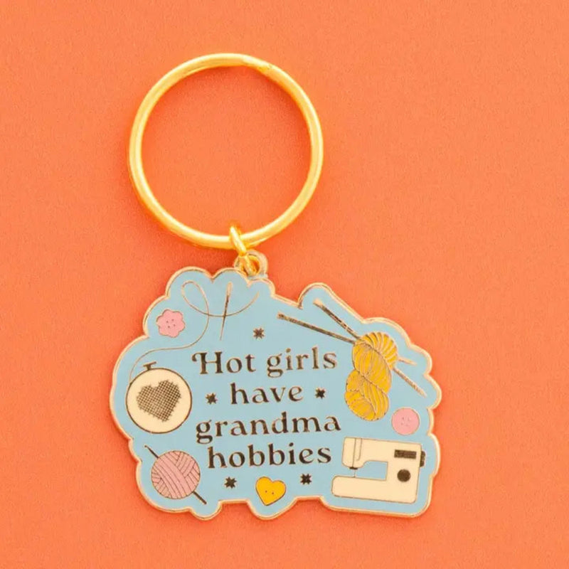 An enamel keychain with a gold-tone split ring and a light blue cloud-shaped charm. The charm features the text 'Hot girls have grandma hobbies', decorated with illustrations, yarn balls, a sewing machine, a cross stitch heart in a hoop and a small yellow heart button.