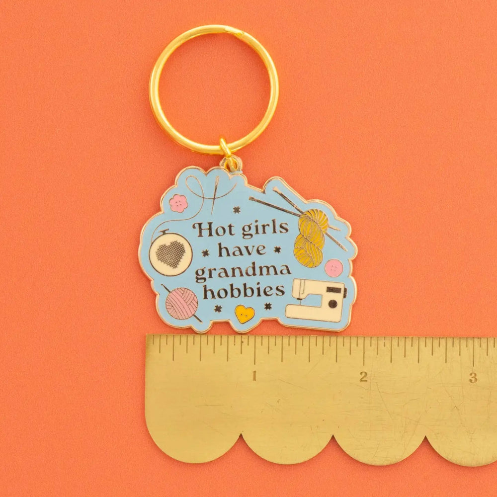 A novelty keychain with a gold-tone split ring and a light blue enamel charm featuring the text 'Hot girls have grandma hobbies' along with craft-themed illustrations, shown next to a gold-colored scalloped edge ruler on a solid orange background.