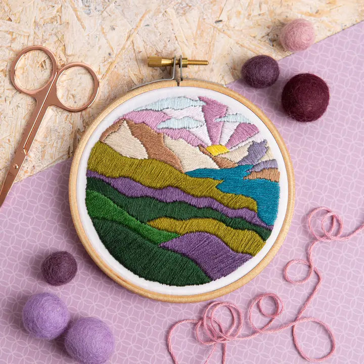 Embroidery hoop with a colorful landscape design on a pink surface with embroidery tools.