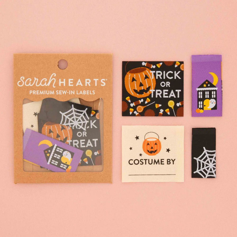 Sarah Hearts premium sew-in Halloween labels displayed on a soft pink background, including a kraft paper package and several individual labels with spooky designs like 'Trick or Treat,' 'Costume by,' spiderwebs, and  a haunted house.