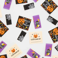 A flat-lay arrangement of Halloween-themed sewing labels on a white background. The labels feature various designs including a black haunted house with a crescent moon and pumpkin on a purple background, a spiderweb on a black background, a jack-o'-lantern with 'TRICK OR TREAT' text, and a cream-colored label that says 'COSTUME BY'.