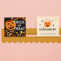 Two Halloween-themed fabric labels  displayed on a gold scalloped ruler against a light pink background. The left tag is black with an orange jack-o'-lantern pail bucket and the words 'TRICK OR TREAT' decorated with candy motifs. The right label is a cream colored label with a jack-o'-lantern pail bucket and the words "Costume By".