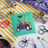 A square green cross-stitch canvas featuring a multi-colored cauldron with bat and cat design in pastel colors, and surrounded by craft tools like scissors, pins, and thread in background.