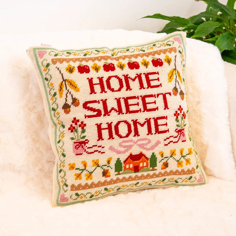 A needlepoint cushion with a the words "Home Sweet Home" in red with a house at the bottom and various floral and nature elements as a border.