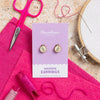 Packaged wooden earrings with pink scissors, thread, and embroidery hoop on a textured surface