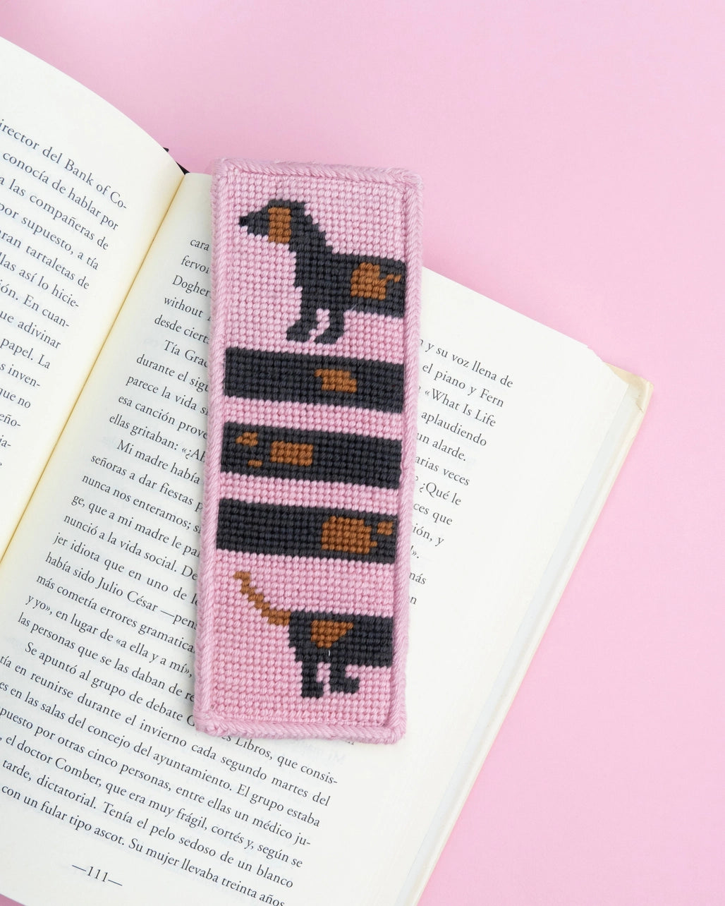 Needlepoint bookmark with a dog design on an open book against a pink background