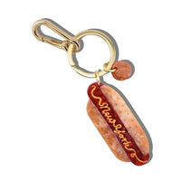 Charm in the shape of a hot dog with text reading "New York" attached to a gold keychain ring.