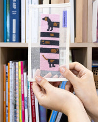 Needlepoint pattern with a dog design held by hands against a bookshelf background