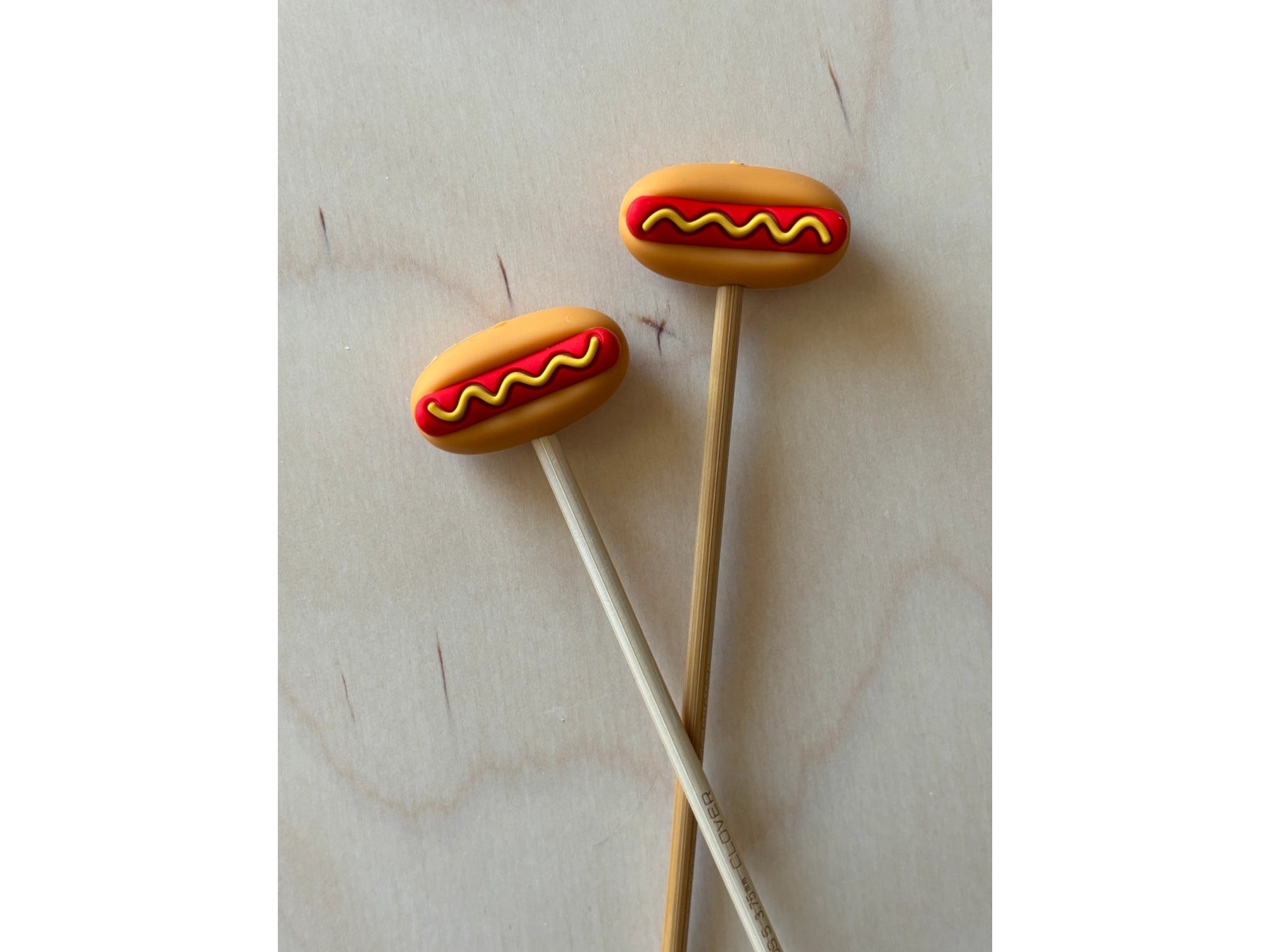 Two hot dog-shaped silicone ends on needles against a light background.