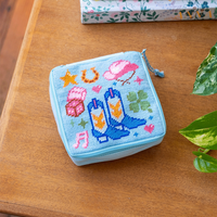 A square zipper pouch with a needlepoint design featuring cowboy boots and a hat, dice and a horseshoe