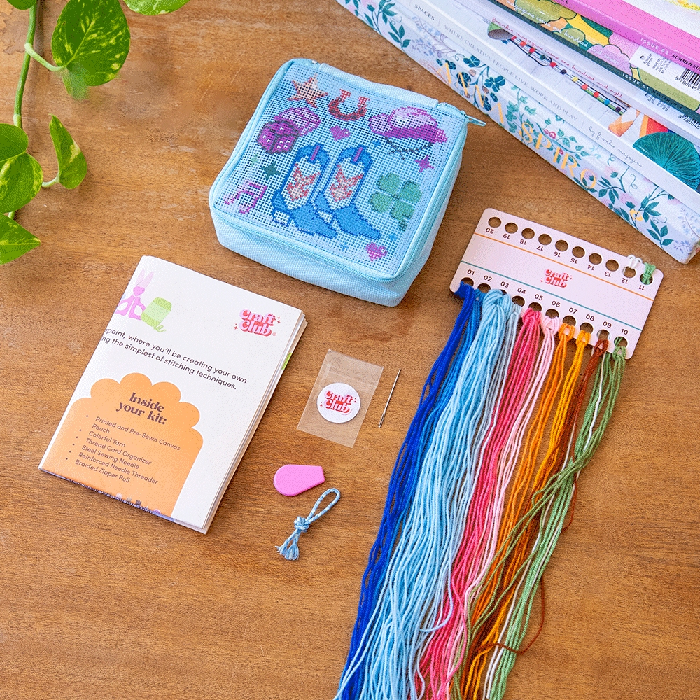 The kit for the needlepoint pouch showing all the included materials like colorful yarn, a needle, pre-sewn pouch and instructions