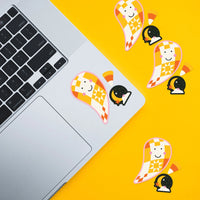A sticker set featuring a cute ghost character with a checkered yellow and orange pattern, a crescent moon, and a book. The sticker is shown applied to a silver laptop on a bright yellow surface, with additional duplicate stickers scattered on the right side.