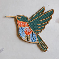 An embroidered iron-on patch of a hummingbird in flight, featuring a teal body and wings with gold embroidery, a bright orange chest, and blue and white patterned details on a light gray background.
