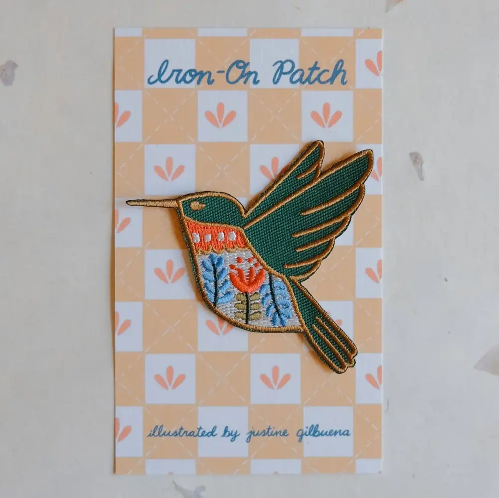 Colorful iron-on patch in the shape of a hummingbird on a piece of cardboard with text reading "Iron-On Patch" and "illustrated by justine gilbuena"