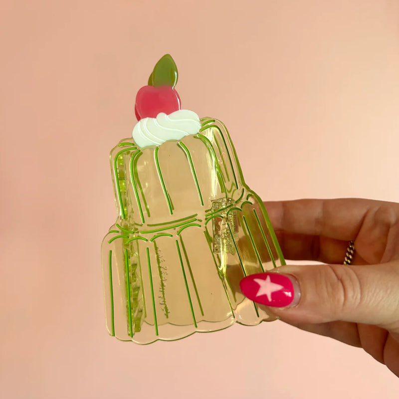Clear green acetate hair claw in the shape of a two-tier jello mold with whipped cream and a cherry on top. Held in a hand with pink star nail art