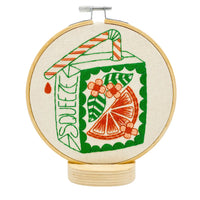 An embroidery hoop mounted on a wooden stand featuring a green and orange juice box design with an orange slice, flowers, and the word "SQUEEZE" on it.