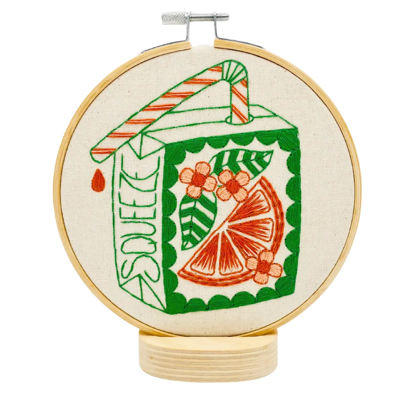 An embroidery hoop mounted on a wooden stand featuring a green and orange juice box design with an orange slice, flowers, and the word "SQUEEZE" on it.