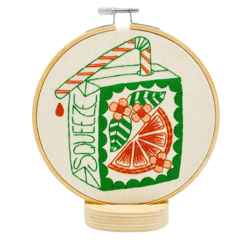 An embroidery hoop mounted on a wooden stand featuring a green and orange juice box design with an orange slice, flowers, and the word "SQUEEZE" on it.