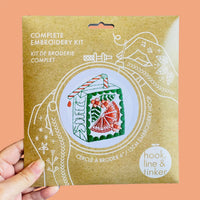 A hand holding a paper package for a complete embroidery kit. The package features white line art of hands holding a hoop and an illustration of an embroidered house with green, red, and orange details.
