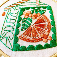Close-up of a hand-embroidered glass cocktail glass design in a hoop, featuring green and orange embroidery on a beige fabric background.