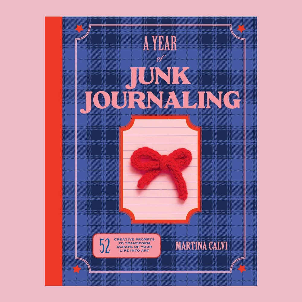 A book titled 'A Year of Junk Journaling' by Martina Calvi, featuring a blue plaid cover with pink lettering and a red yarn bow illustration, set against a solid pink background.