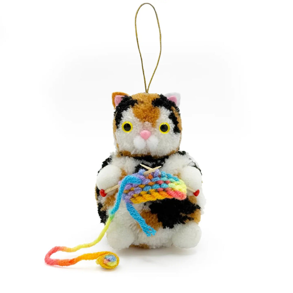 A plush calico cat hanging ornament holding a small rainbow-colored knitted ball, with a gold hanging loop at the top, photographed on a white background.