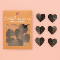 Sarah Hearts Premium Buttons in a kraft paper packaging, shown next to six heart-shaped buttons in black and gray wood grain on a solid pink background.