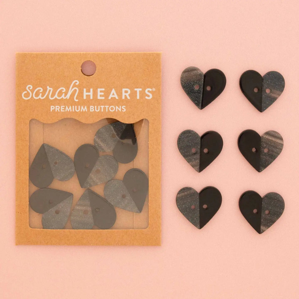Sarah Hearts Premium Buttons in a kraft paper packaging, shown next to six heart-shaped buttons in black and gray wood grain on a solid pink background.