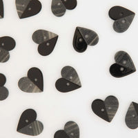 Heart-shaped buttons scattered on a white background, featuring a split design with one half solid glossy black and the other half a textured sparkly grey pattern.