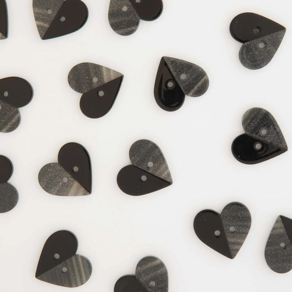 Heart-shaped buttons scattered on a white background, featuring a split design with one half solid glossy black and the other half a textured sparkly grey pattern.