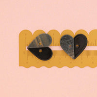 Two heart-shaped buttons,half black and half grey with silver glitter, shown on a yellow scalloped ruler against a light pink background.