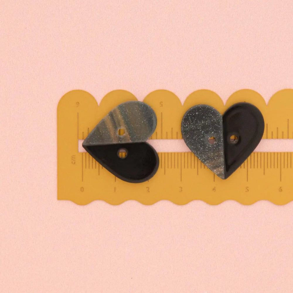 Two heart-shaped buttons,half black and half grey with silver glitter, shown on a yellow scalloped ruler against a light pink background.