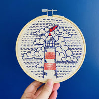 A hand holds a round wooden embroidery hoop containing a stitched design of a lighthouse with red and white stripes, blue clouds, and blue wave-like lines on a cream fabric, set against a solid blue background.