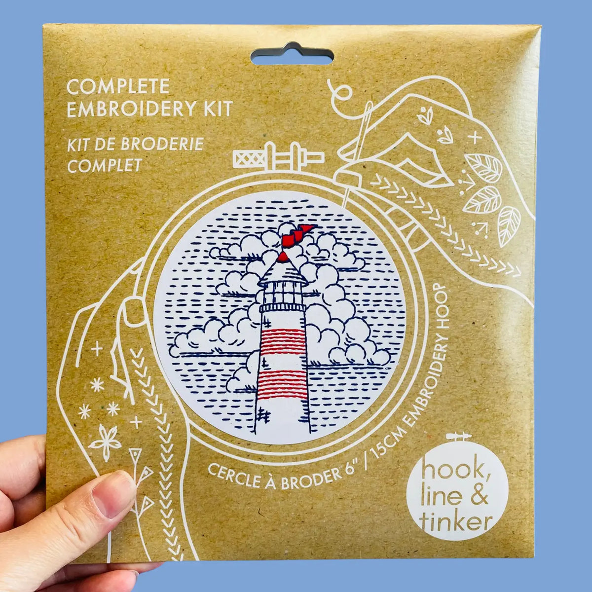 A hand holding a paper package for a complete embroidery kit. The package features a lighthouse illustration in blue and red, with white text reading 'COMPLETE EMBROIDERY KIT' and a 'hook, line & tinker' logo.