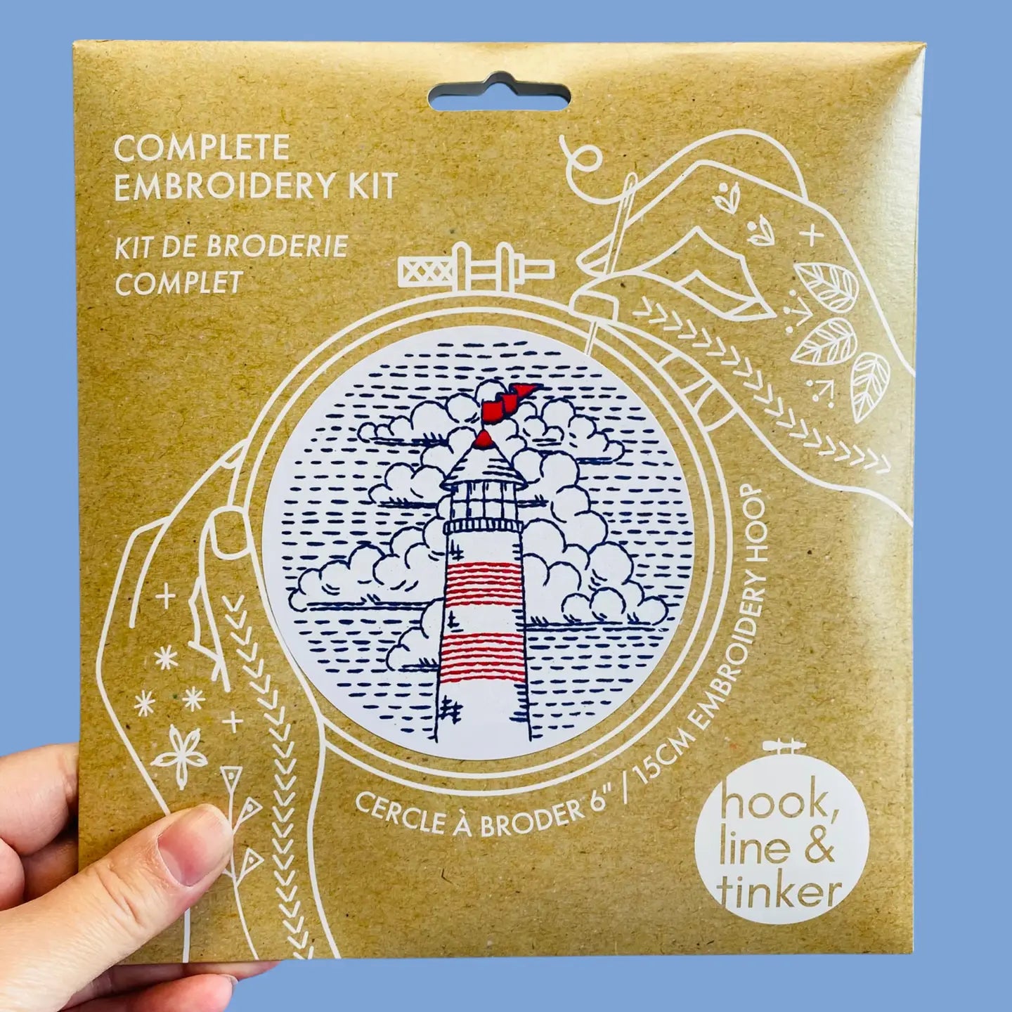 A hand holding a paper package for a complete embroidery kit. The package features a lighthouse illustration in blue and red, with white text reading 'COMPLETE EMBROIDERY KIT' and a 'hook, line & tinker' logo.