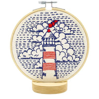 A circular embroidery hoop containing a stitched lighthouse design with blue clouds and dotted water, on a white background.