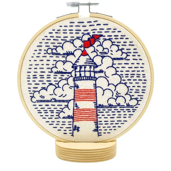 A circular embroidery hoop containing a stitched lighthouse design with blue clouds and dotted water, on a white background.