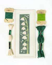 Needlepoint kit with a pattern of white flowers on a green background, surrounded by green and beige thread skeins.