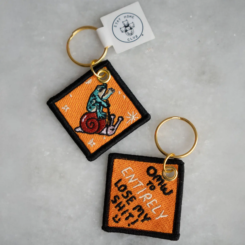 Two square keychains with embroidered frog & snail. Text on one side read on a gray surface, 'OMW to ENTIRELY lose my shit'