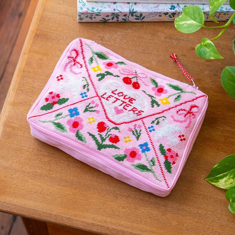 A rectangular zipper pouch with a needlepoint design resembling a pink envelope with the text 'love letters'